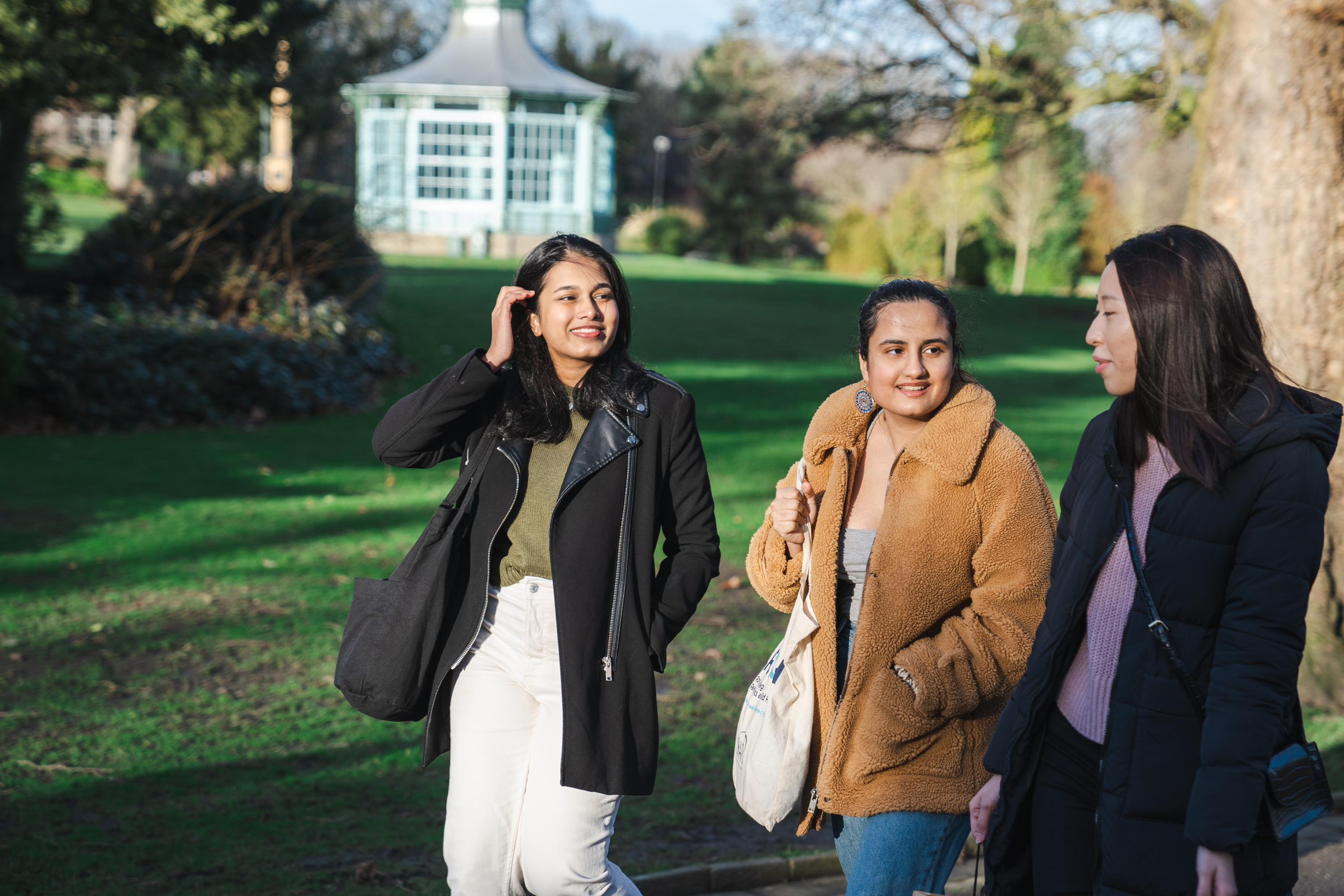 Three people walking together along a paved path in a park with green grass and trees. They are dressed in warm, casual clothing, including coats and jackets, and carrying bags. In the background, there is a light blue gazebo surrounded by greenery, and the scene is lit by natural daylight.