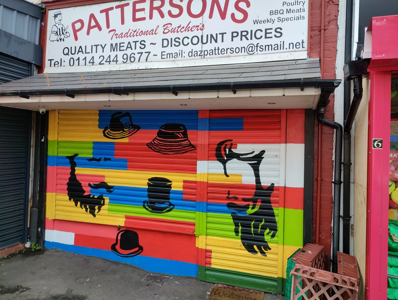 A butcher shop with a closed shutter painted in bright horizontal stripes of red, yellow, green, blue, and white. The mural features black and red silhouettes of various hats and a stylized profile of a man with a beard and hat. Above the shutter is a sign reading “Pattersons Traditional Butchers” with contact details and offers for quality meats and discount prices.