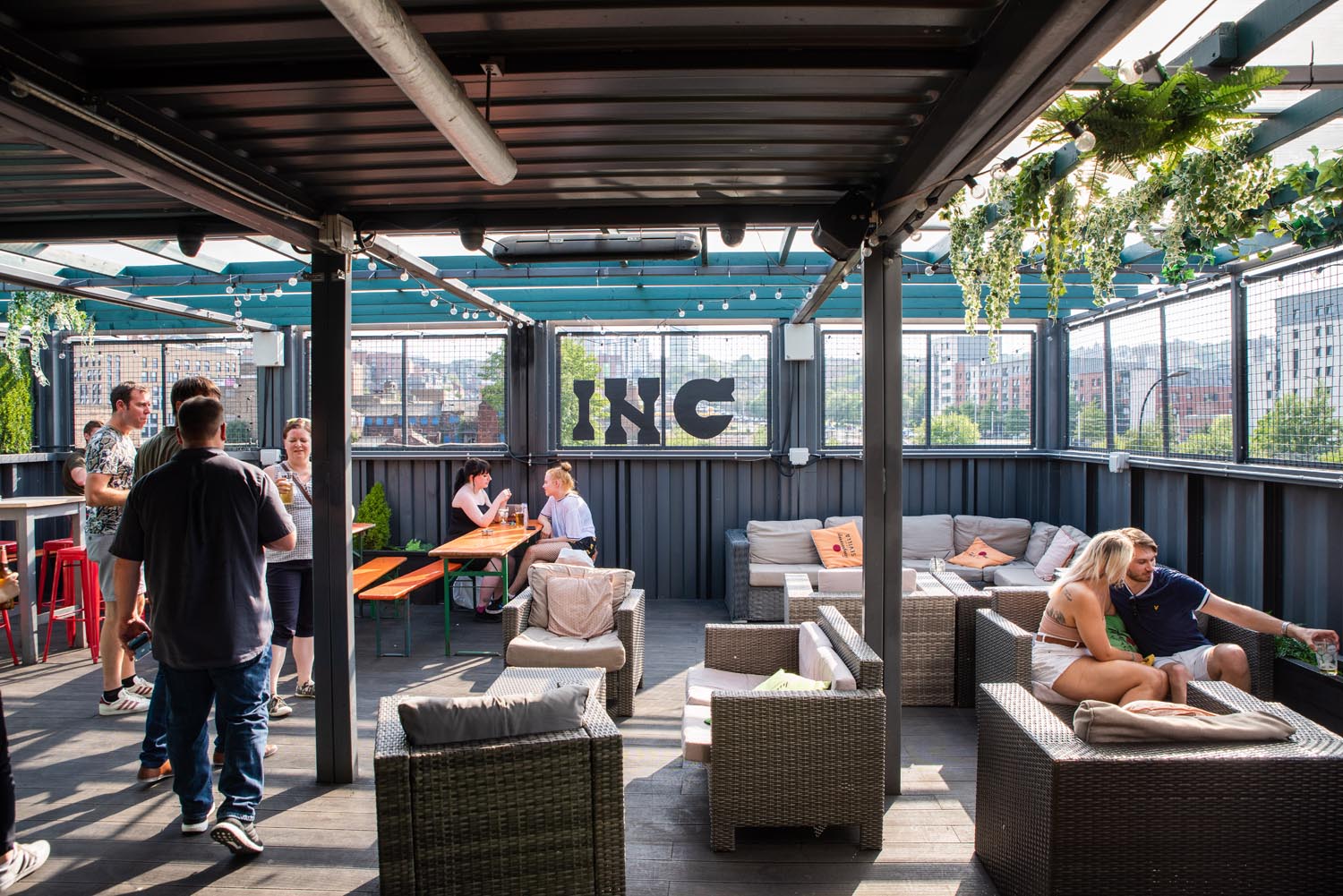 INC Sheffield - Outdoor rooftop bar with a covered seating area featuring wicker sofas with cushions, wooden tables, and string lights overhead. Green plants hang from the ceiling, and the space offers views of city buildings through mesh fencing. Several people are standing and sitting, enjoying drinks and conversation.