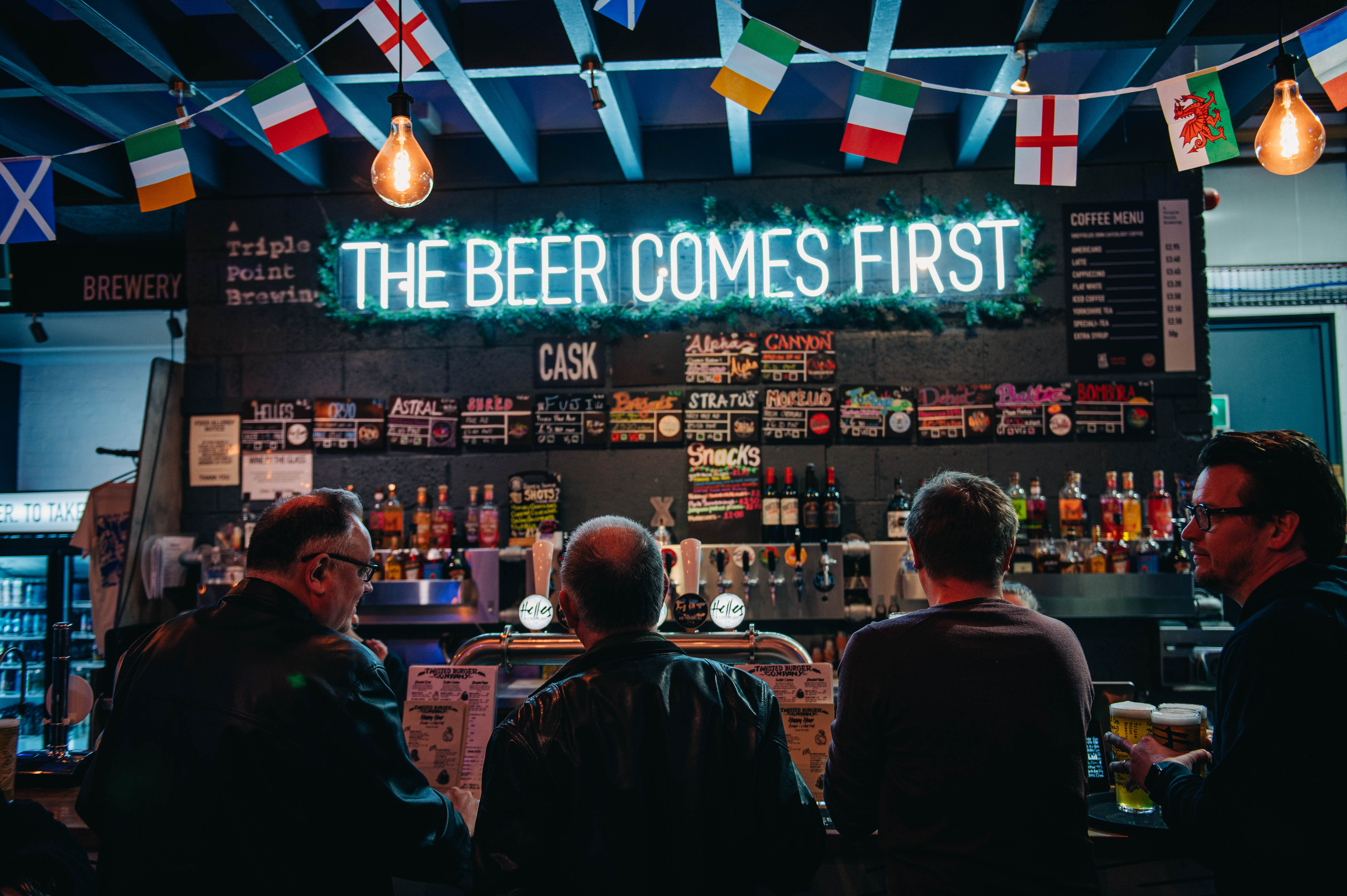 Triple Point Brewery bar with blue neon sign above that reads 'The Beer Comes First' and international flag bunting. Four people are stood waiting at the bar.