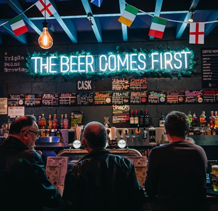 Triple Point Brewery bar with blue neon sign above that reads 'The Beer Comes First' and international flag bunting. Four people are stood waiting at the bar.