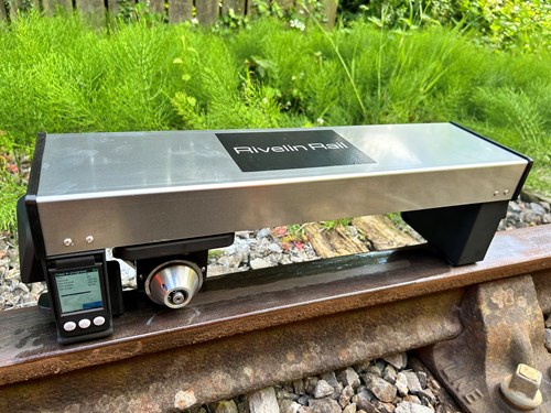 Metallic device labeled ‘Rivelin Rail’ positioned on a railway track. The device has a rectangular body with a digital display and control buttons on one end, and a circular component underneath. Surrounding the track are gravel stones and green vegetation in the background.