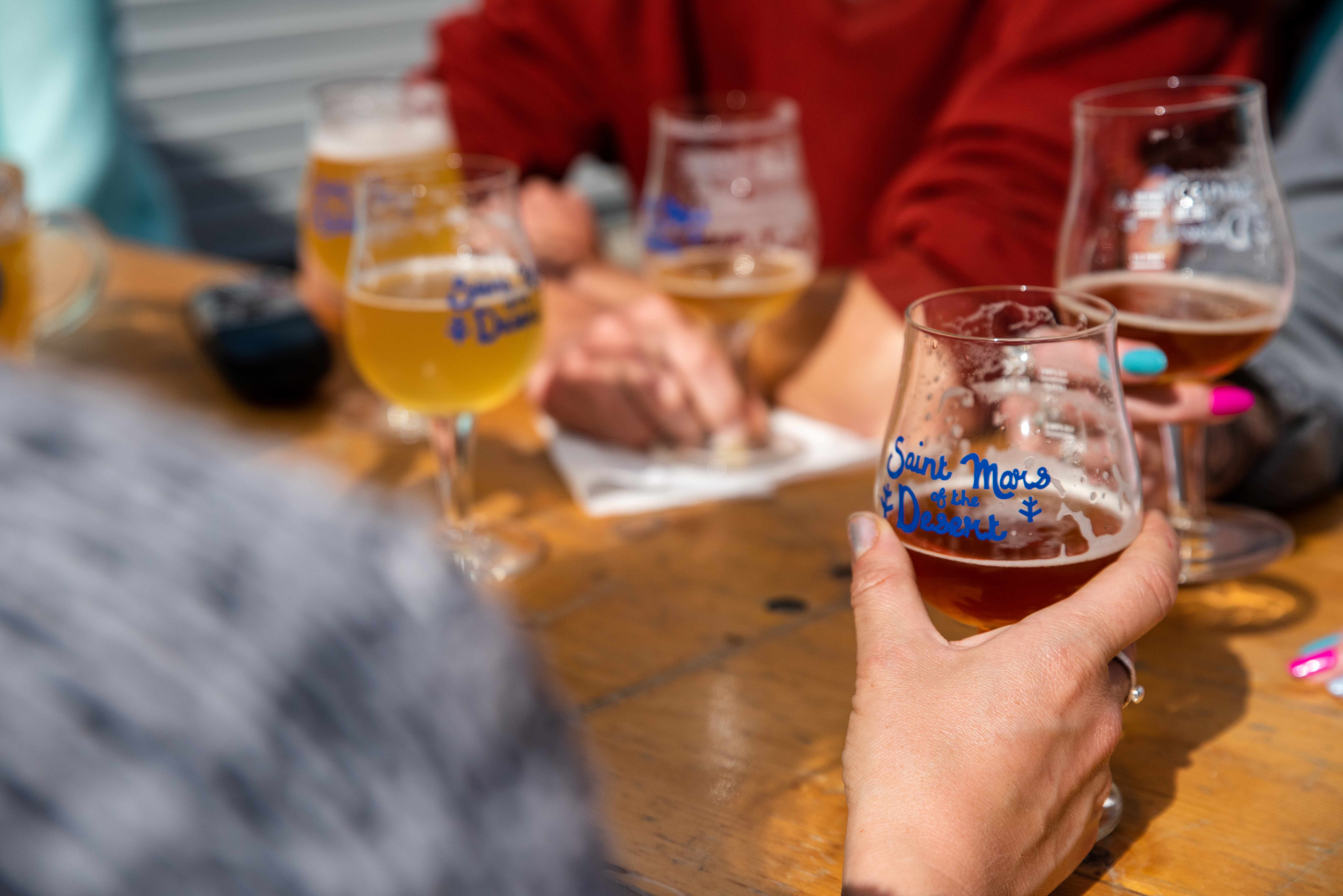 Close-up of several people sitting around a wooden table with multiple glasses of craft beer. One hand in the foreground holds a glass labeled ‘Saint Mars of the Desert.’ Other glasses with similar labels and varying shades of beer are visible, along with a notepad and pen on the table.