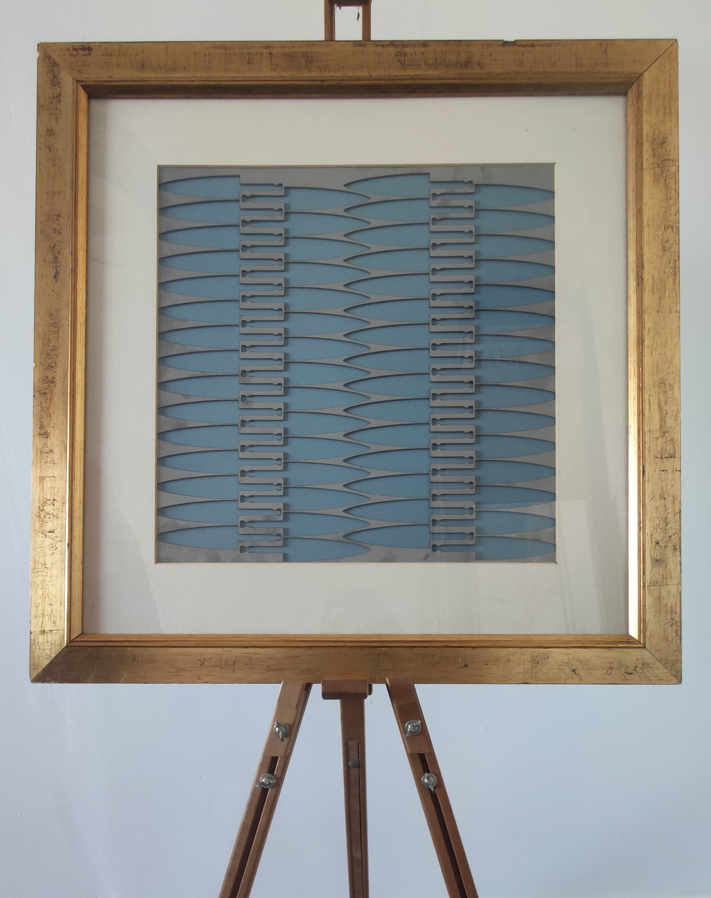 Framed artwork with a gold frame showcasing a blue background and metallic cut-out shapes arranged in vertical rows, displayed on a wooden easel.