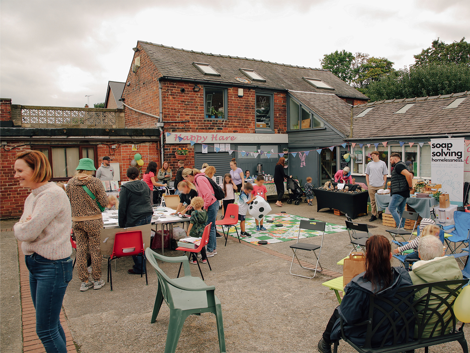 Outdoor community event in a courtyard with people gathered around tables and chairs. Activities include arts and crafts on a table, a large board game with a giant dice on the ground, and stalls selling items. The setting features red brick buildings with signs reading ‘Happy Hare’ and ‘Soap Solving Homelessness.’ Balloons and decorations add a festive atmosphere.