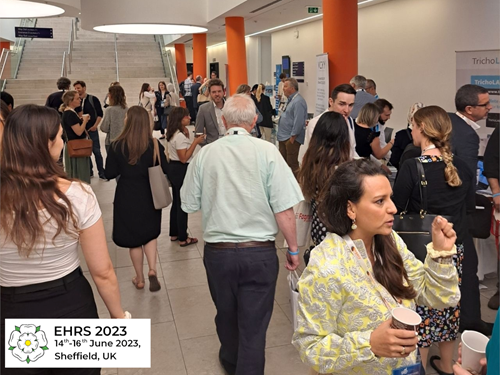 A busy conference networking area with attendees standing and conversing near exhibition tables. The setting features a wide hallway with bright lighting, orange accent columns, and a staircase in the background. People are holding cups, brochures, and bags, and some are gathered around informational displays. A sign in the bottom-left corner reads “EHRS 2023, 14th–16th June 2023, Sheffield, UK.”