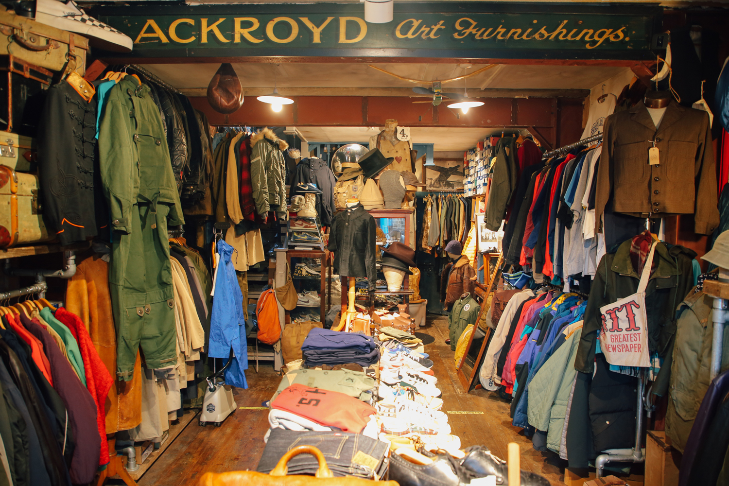 The interior of a clothes shop. Both walls are filled with hanging cloths. In the centre of the shop is a long table filled with folded clothes. 