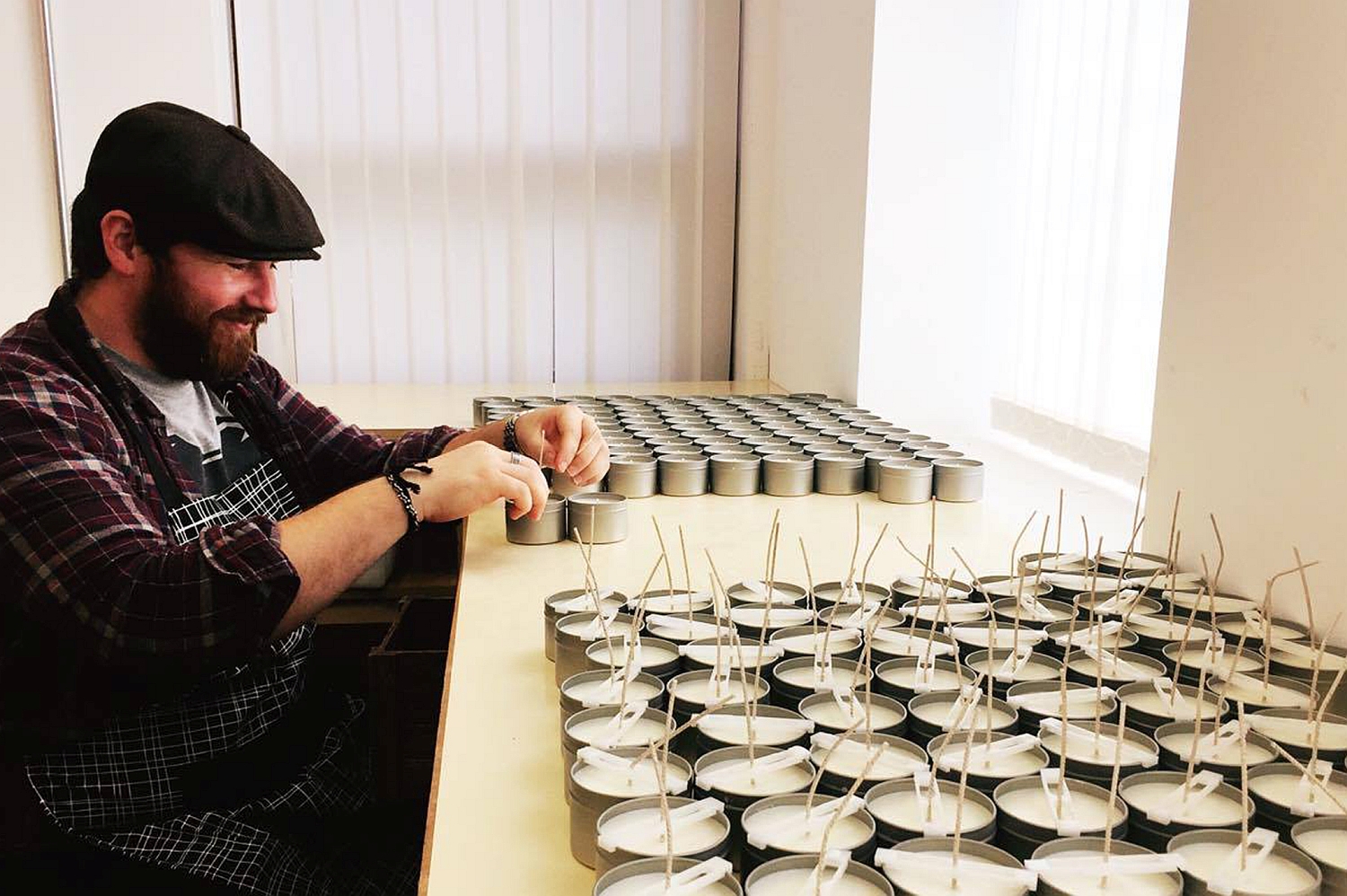 A bearded man, wearing a flat cap and an apron, sits at a desk adding wicks to candles.