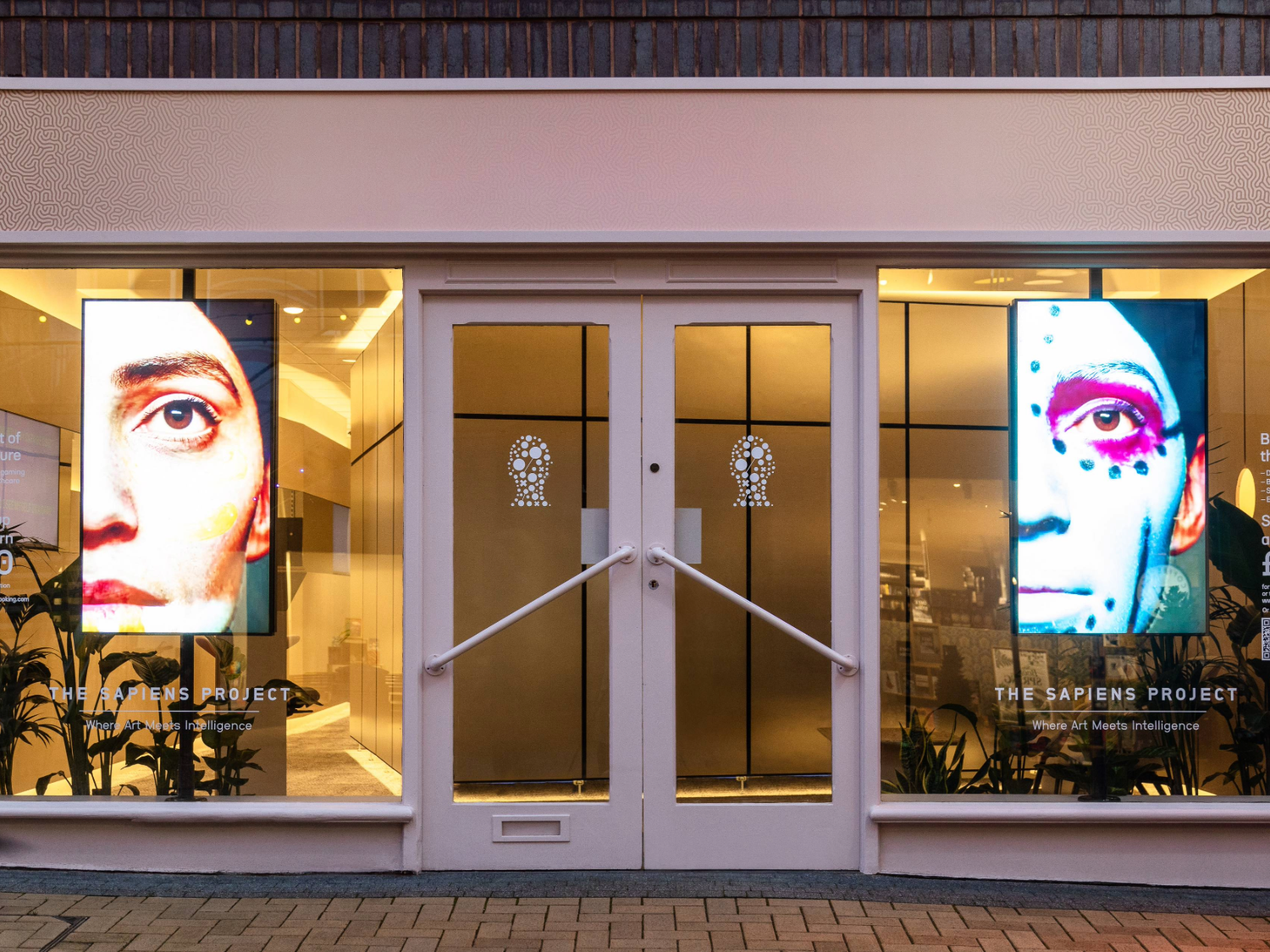 Storefront with two large illuminated digital screens displaying close-up portraits of faces with artistic designs, part of an installation called ‘The Sapiens Project.’ The entrance features double glass doors with decorative patterns, and the interior is warmly lit.