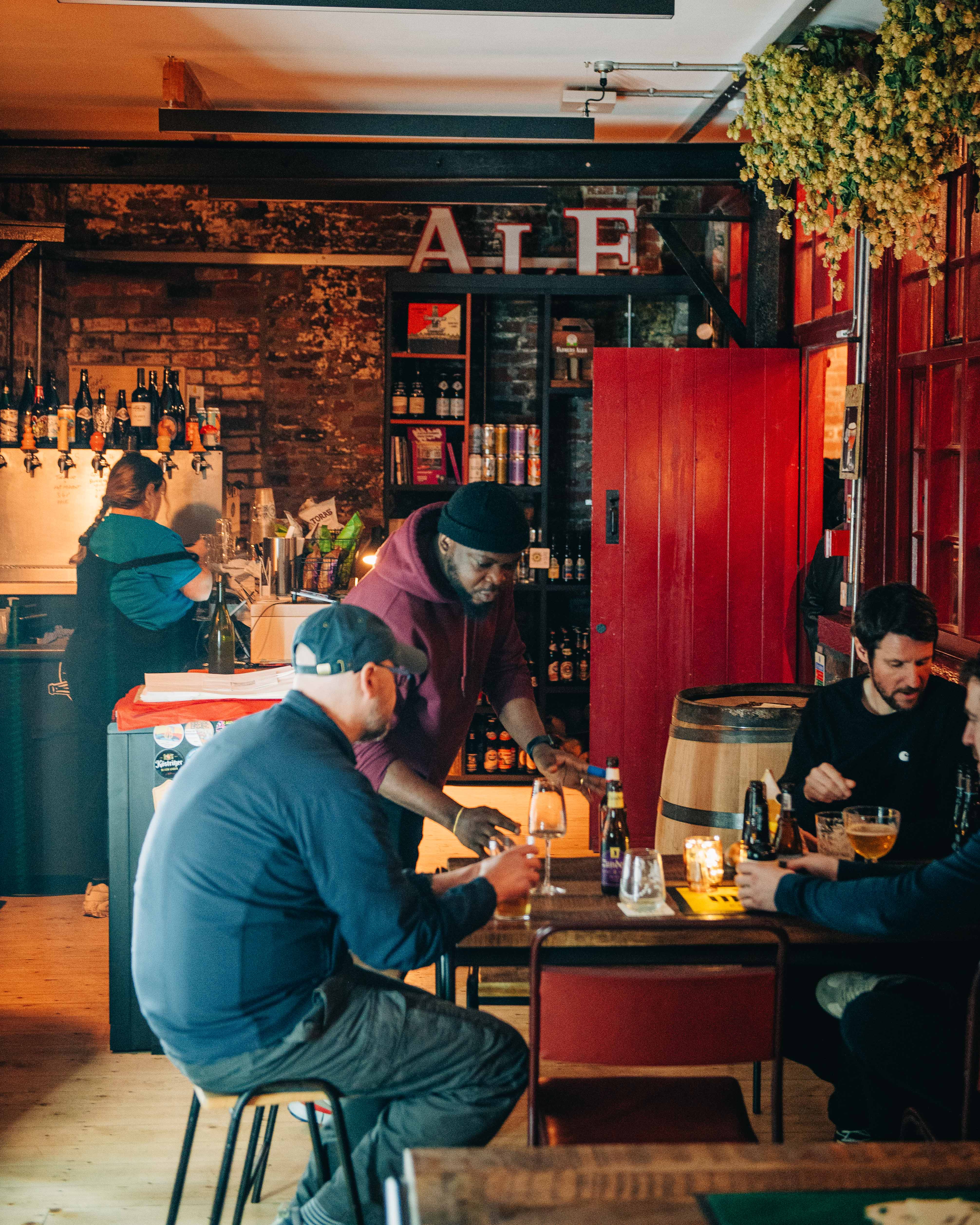 Three men are sat drinking in Hop Hideout, with a male bartender putting a drink down on their table. A female bartender is in the background at the bar on the left, with a shelving unit full of their stock to the right by the entrance.