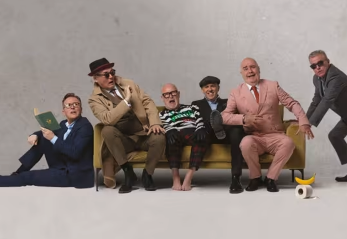 The band Madness all piled on to and around a yellow sofa.