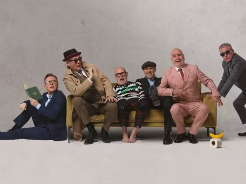 The band Madness all piled on to and around a yellow sofa.