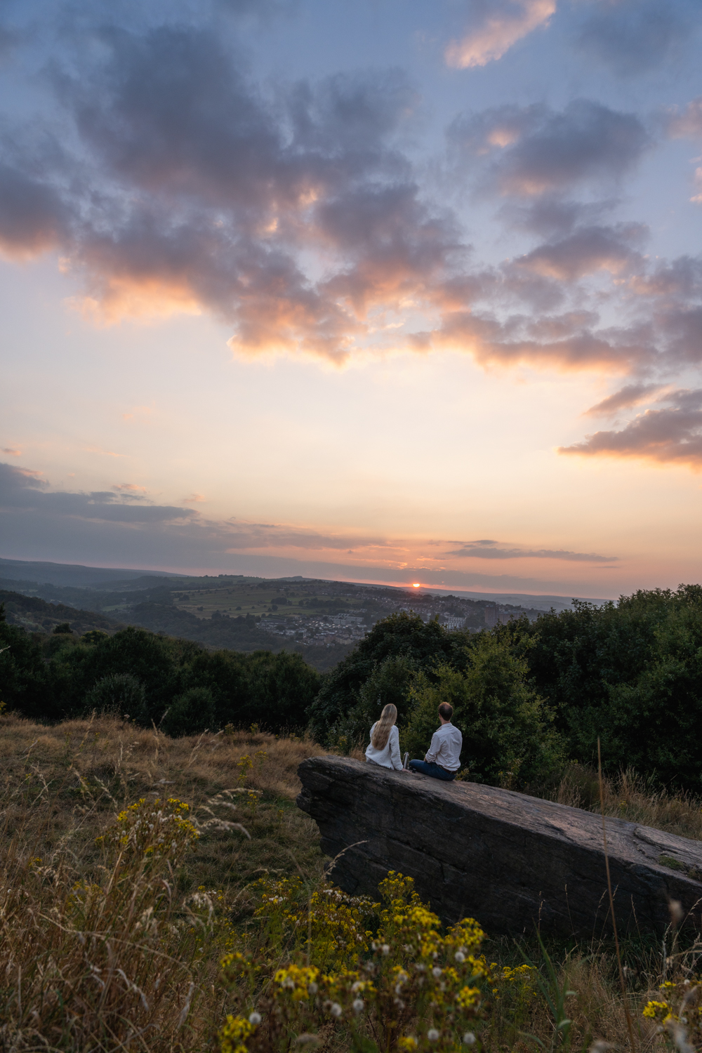 Two people sitting on a large rock in a grassy hillside area, overlooking a scenic valley with rolling green hills and a village in the distance. The sun is setting on the horizon, casting a warm golden light across the landscape, with trees and wildflowers in the foreground.