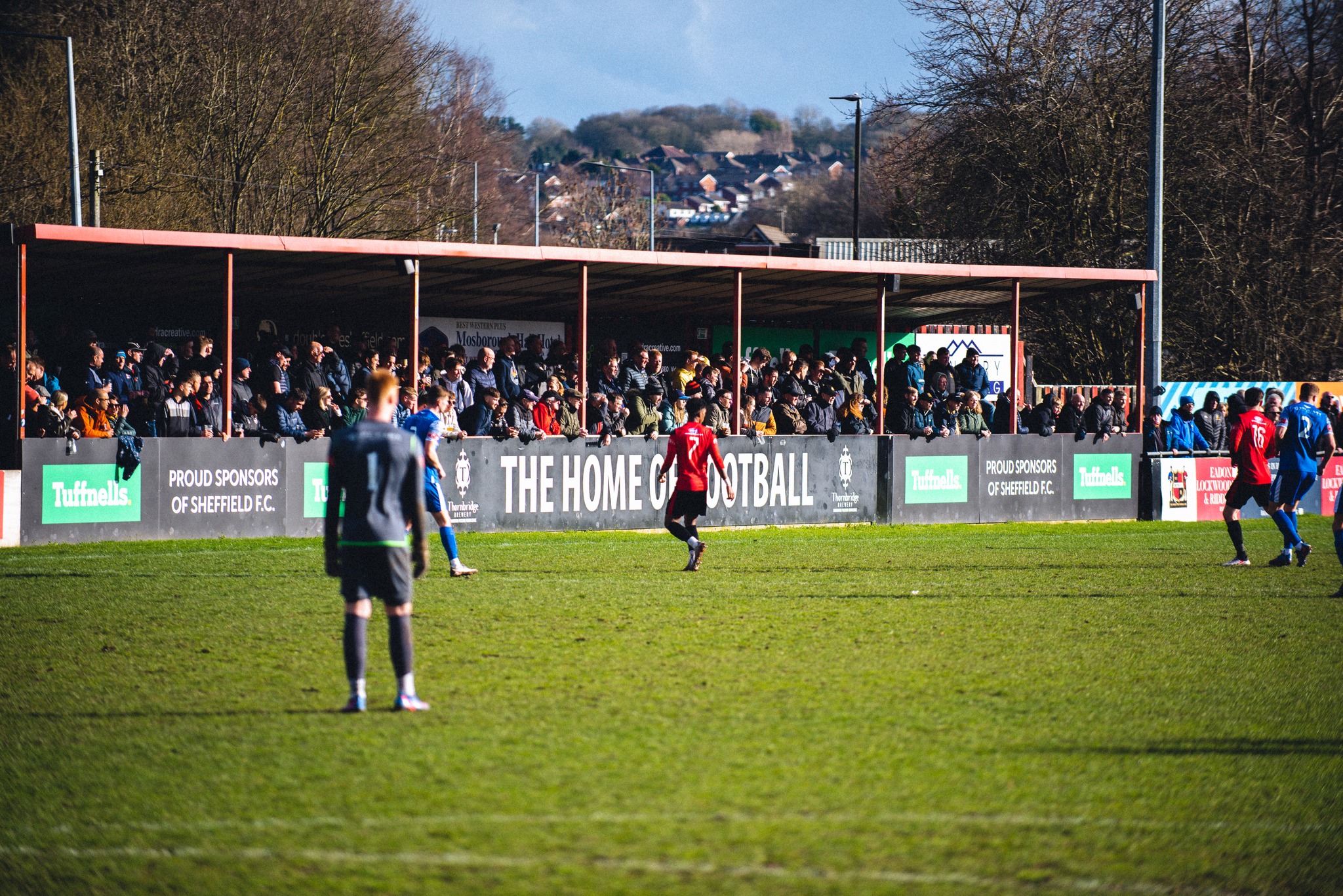 Outdoor football match at a small stadium with players on the pitch and a crowd of spectators standing under a covered stand. Advertising boards along the stand display text including ‘The Home of Football’ and sponsor logos. Trees and houses are visible in the background.