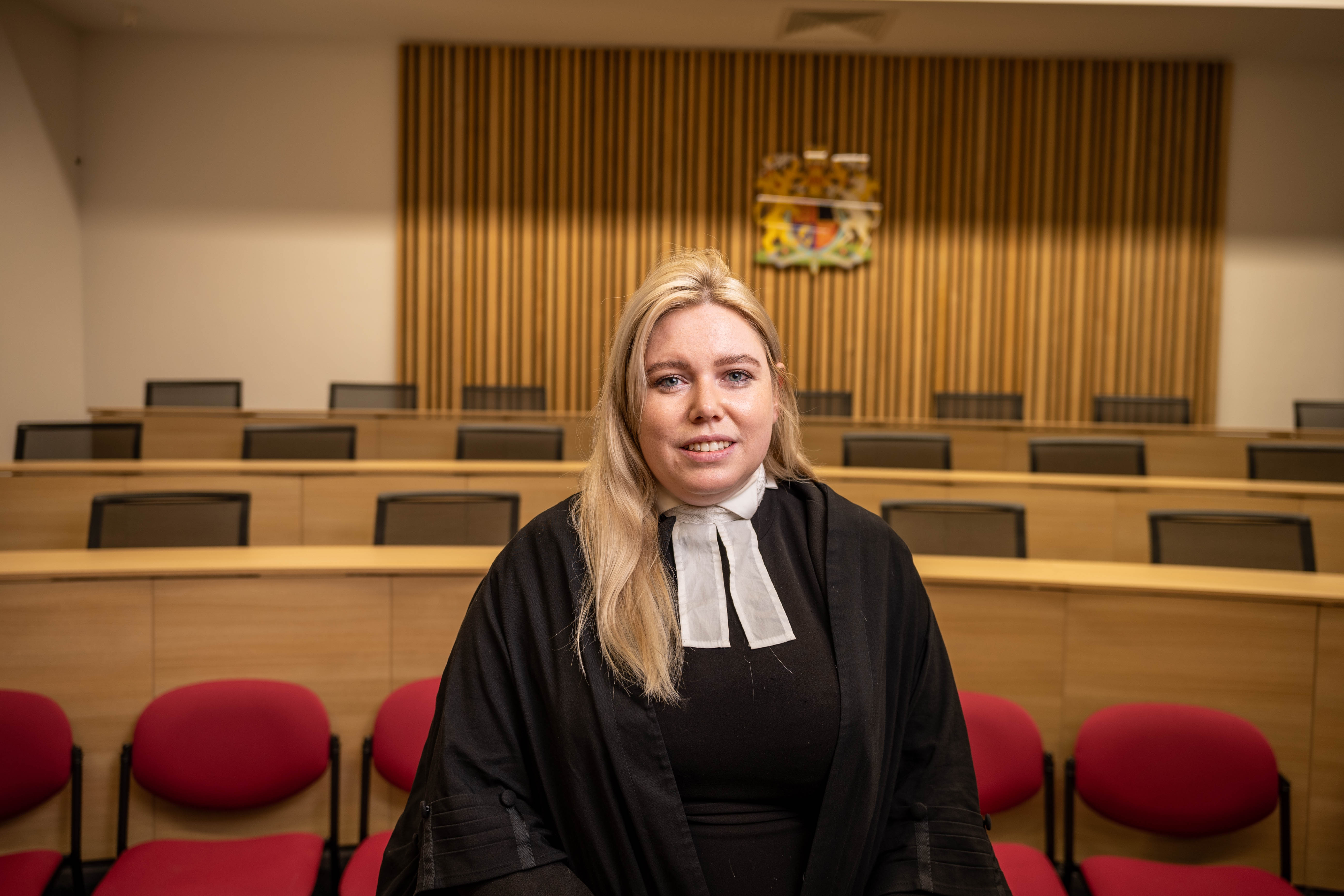 A person wearing a black robe and white neck bands is standing in a courtroom setting. Behind them are rows of red chairs and wooden benches, with a wooden panel wall featuring a coat of arms at the centre. The lighting is warm, and the room has a formal, official atmosphere.