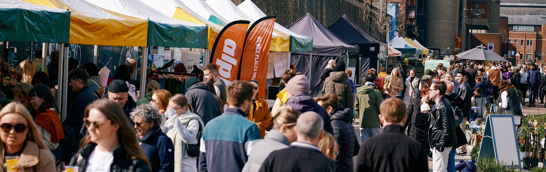 An open air market in a city centre. There are lots of people walking up and down and browsing the stalls.