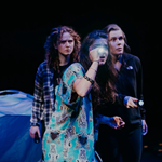 Three women, onstage as part of a play, are looking worried. Two of them are holding torches. Behind them is a blue tent.