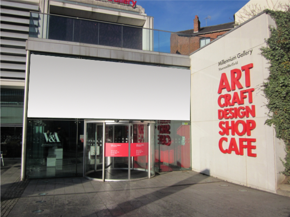 Entrance to the Millennium Gallery in Sheffield, featuring a modern glass facade with a revolving door. Large red text on the right wall reads “ART CRAFT DESIGN SHOP CAFE.” A blank white sign is displayed above the entrance, and the building has a clean, contemporary design with concrete and glass elements.