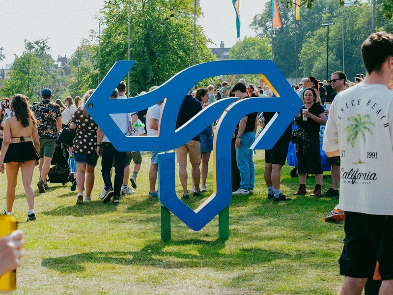 A giant sculpture of the Tramlines festival logo stands on the grass as a crowds of festivalgoers walk by