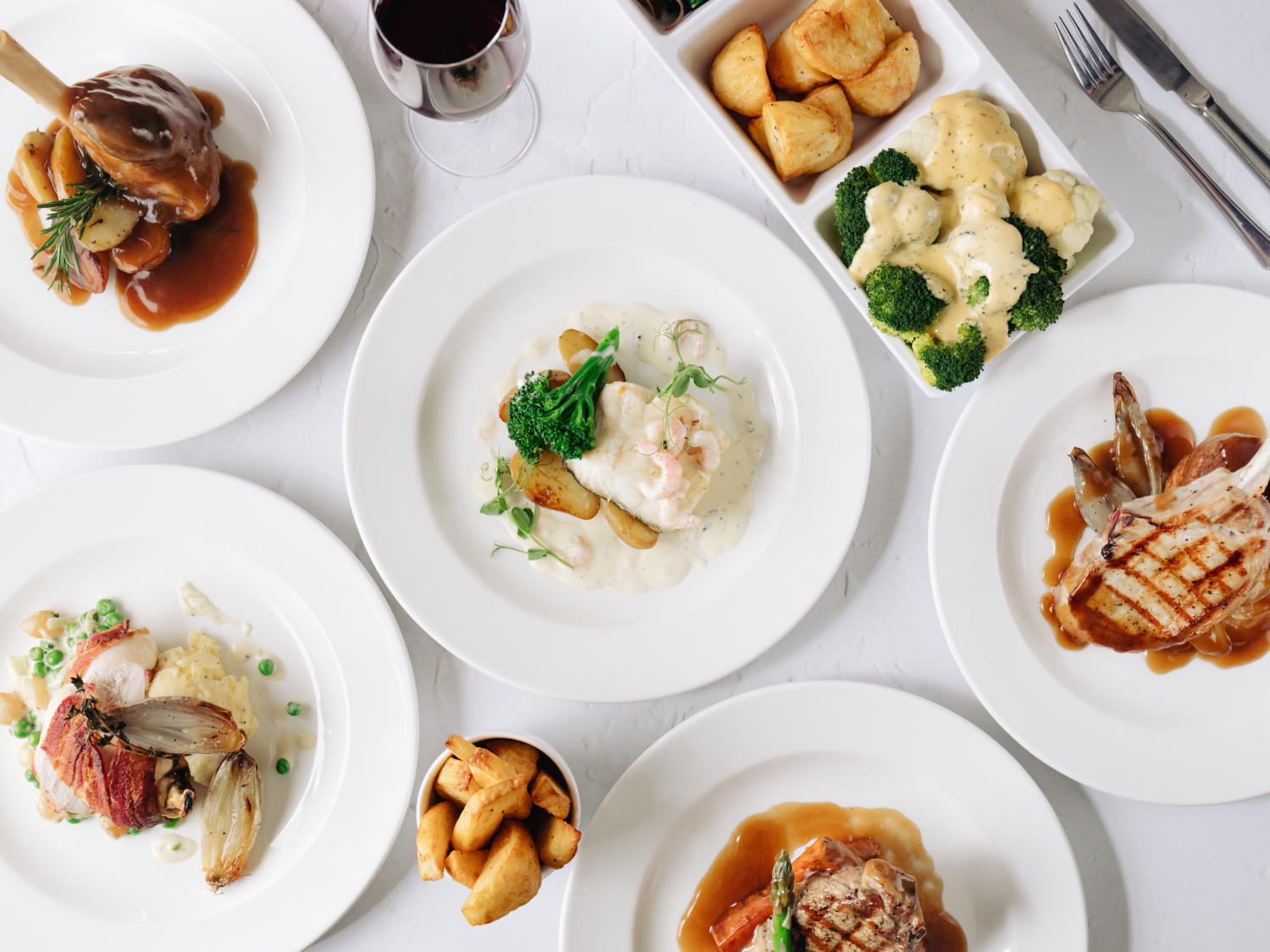 An overhead view of a table laid out with 7 plates of food and a glass of red wine.