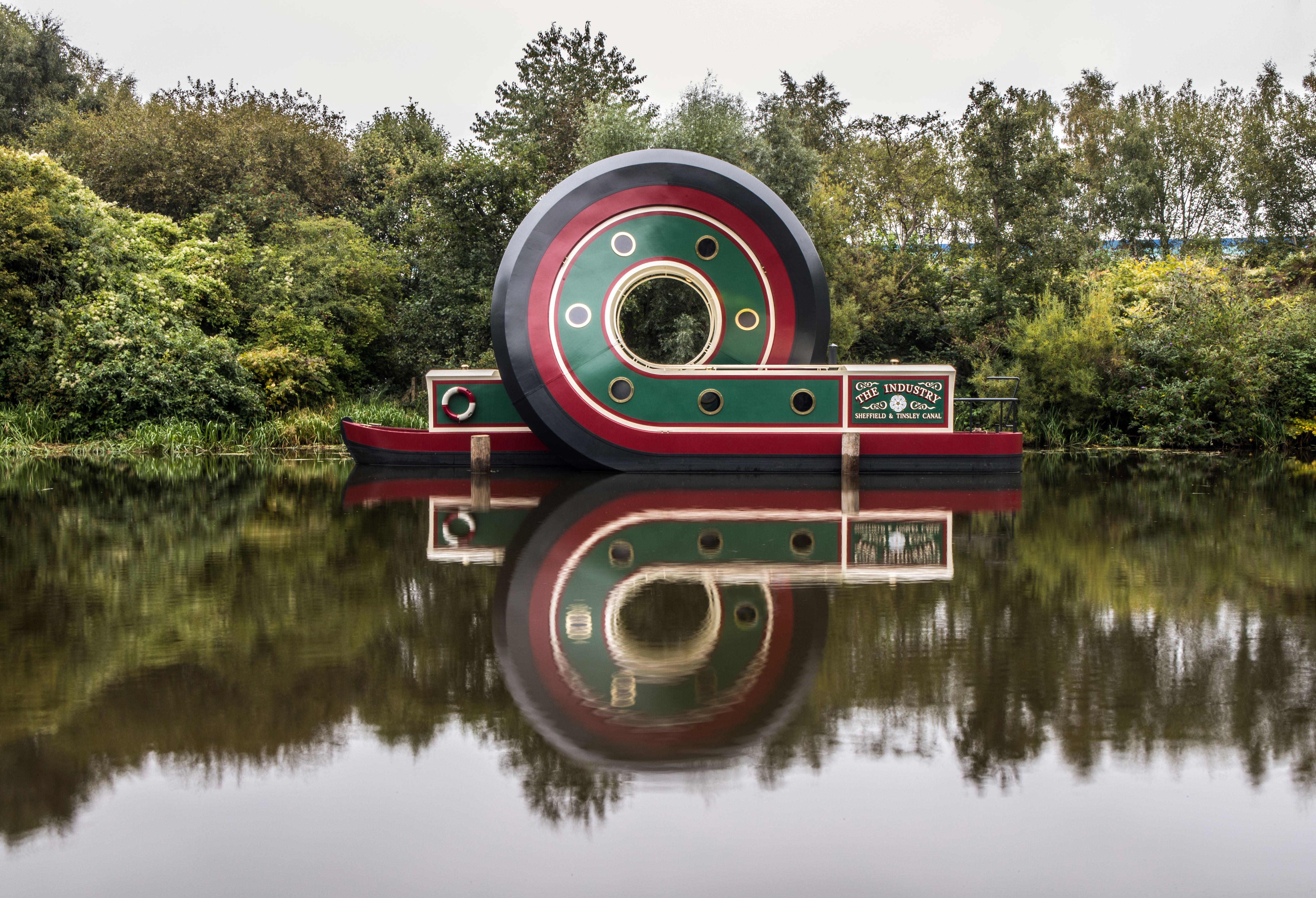 Large circular sculpture resembling a stylized boat with green, red, and black colors floating on a calm river, creating a clear reflection in the water. Dense green trees and shrubs form the background.