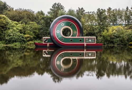 Large circular sculpture resembling a stylized boat with green, red, and black colors floating on a calm river, creating a clear reflection in the water. Dense green trees and shrubs form the background.