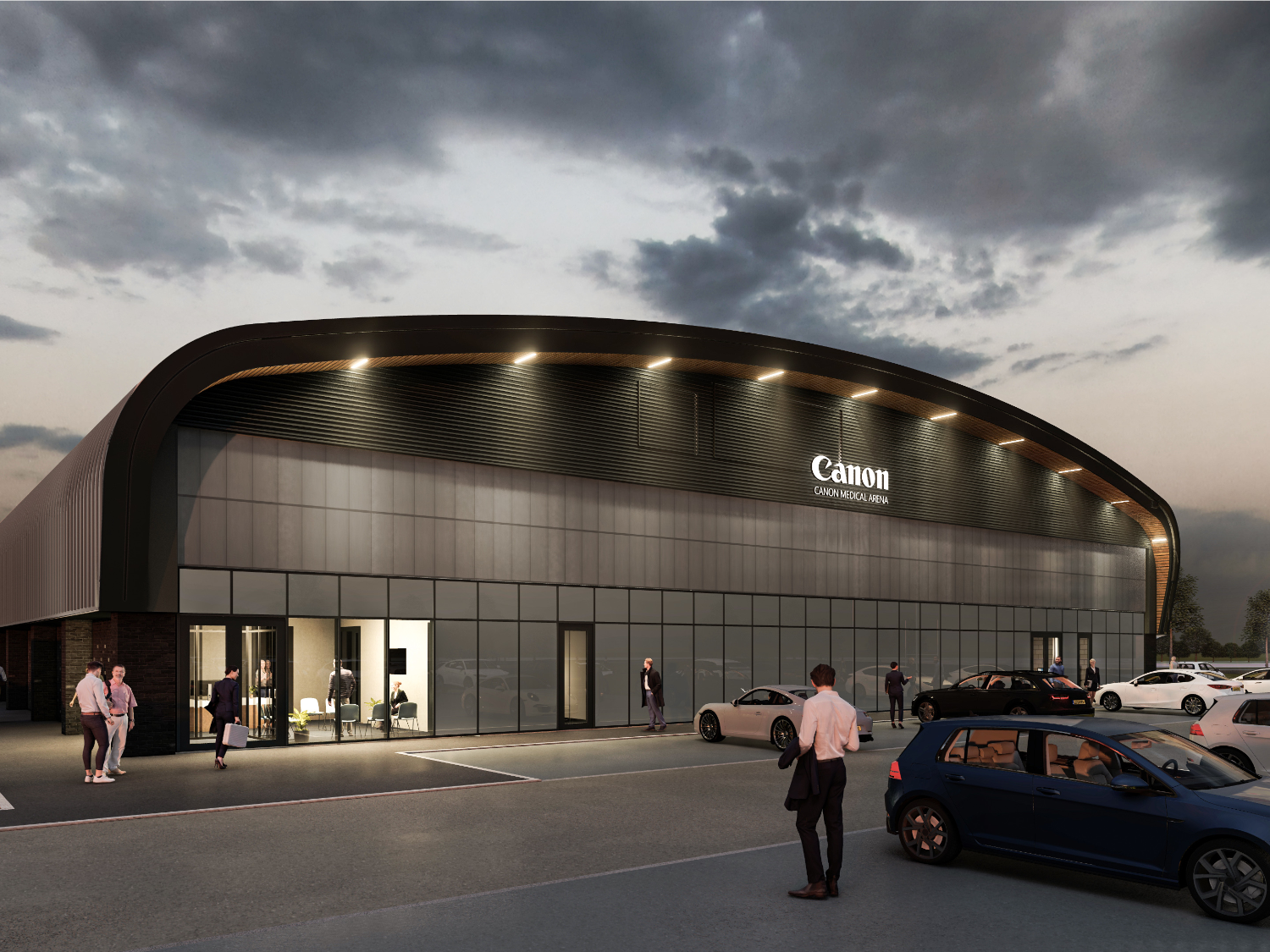 The exterior of the Canon Medical Arena, a building made of glass and steel with a gently curved roof, as seen from the car park in the evening.