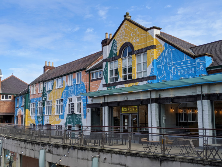 Row of connected buildings with pitched roofs featuring a large, colorful mural across the façade. The artwork includes abstract shapes and line drawings in blue, yellow, green, and orange. A sign reading ‘FOOLS GOLD’ is above the entrance to a ground-floor venue, and there is an outdoor seating area with tables and chairs along a raised walkway.