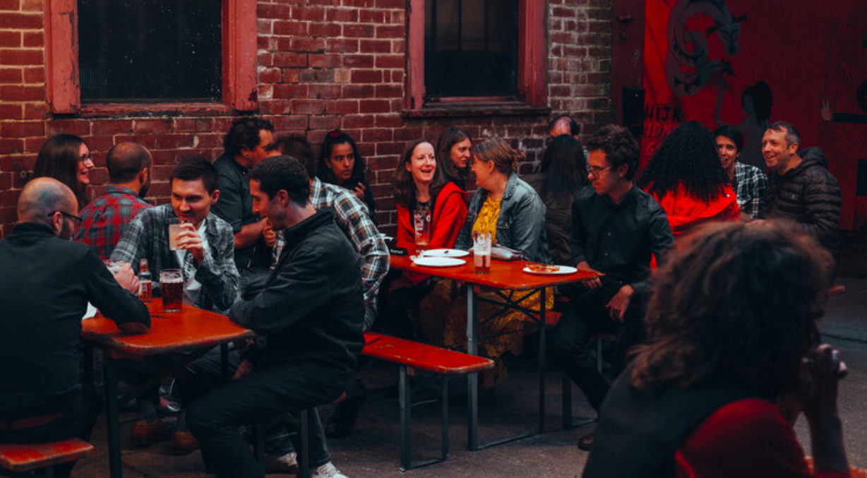 A group of people are sat at tables outside, at night, chatting and having a drink. 