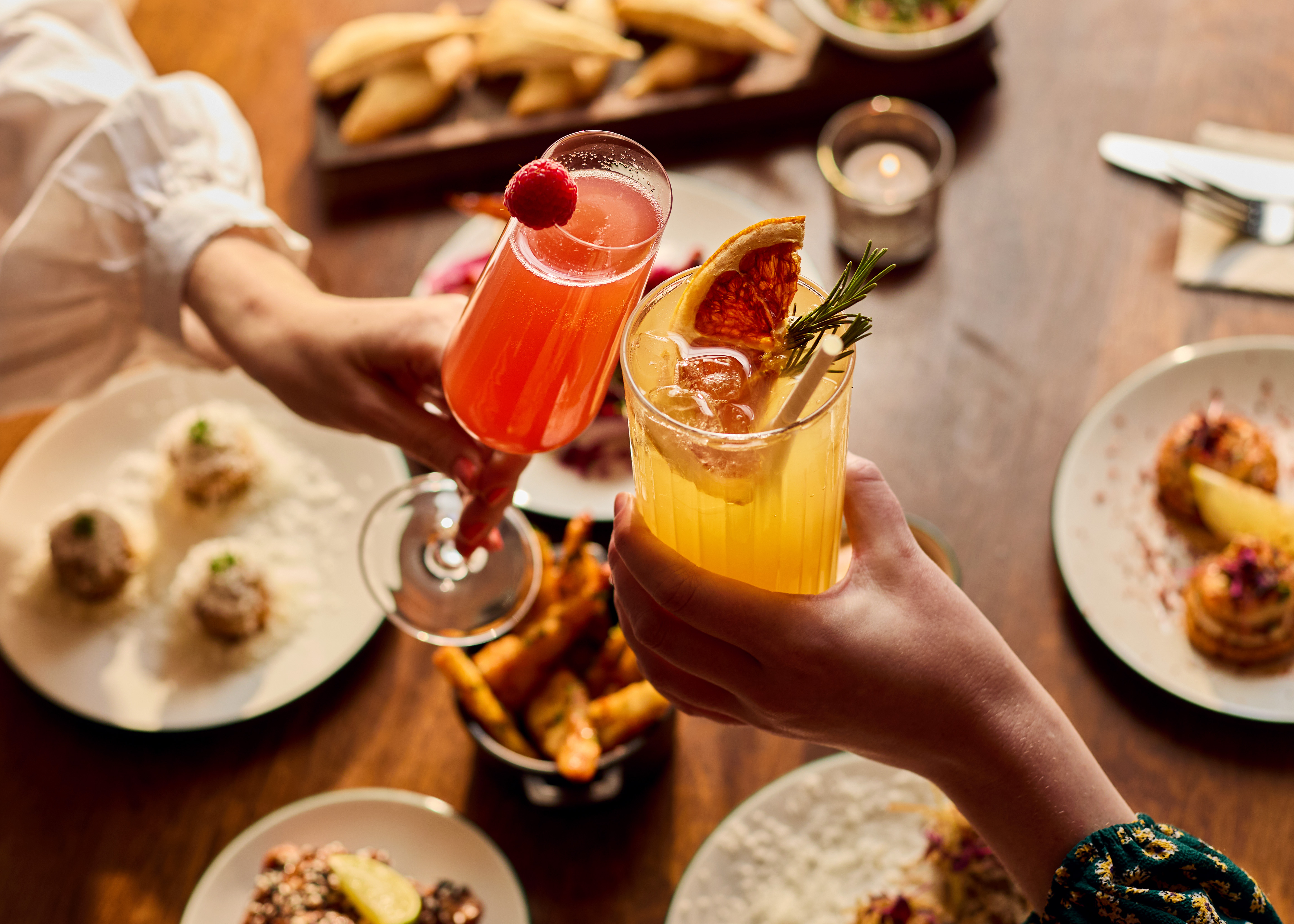 Two hands hold colourful cocktails above a table filled with delicious food.