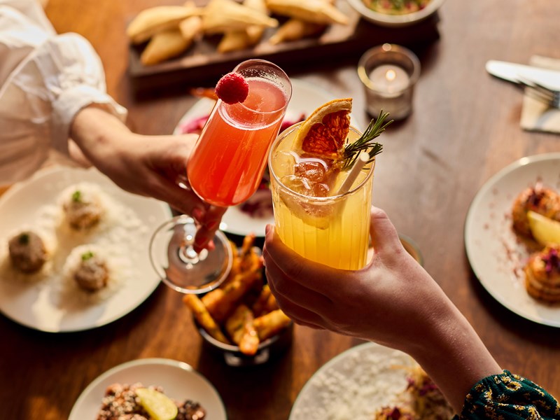Two hands hold colourful cocktails above a table filled with delicious food.