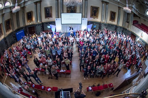 Large group of people gathered inside a grand hall with chandeliers, high ceilings, and framed portraits on the walls. The audience is standing and facing the stage, where a speaker is positioned in front of a screen displaying ‘ITI Conference.’ Rows of red chairs are visible in the foreground.