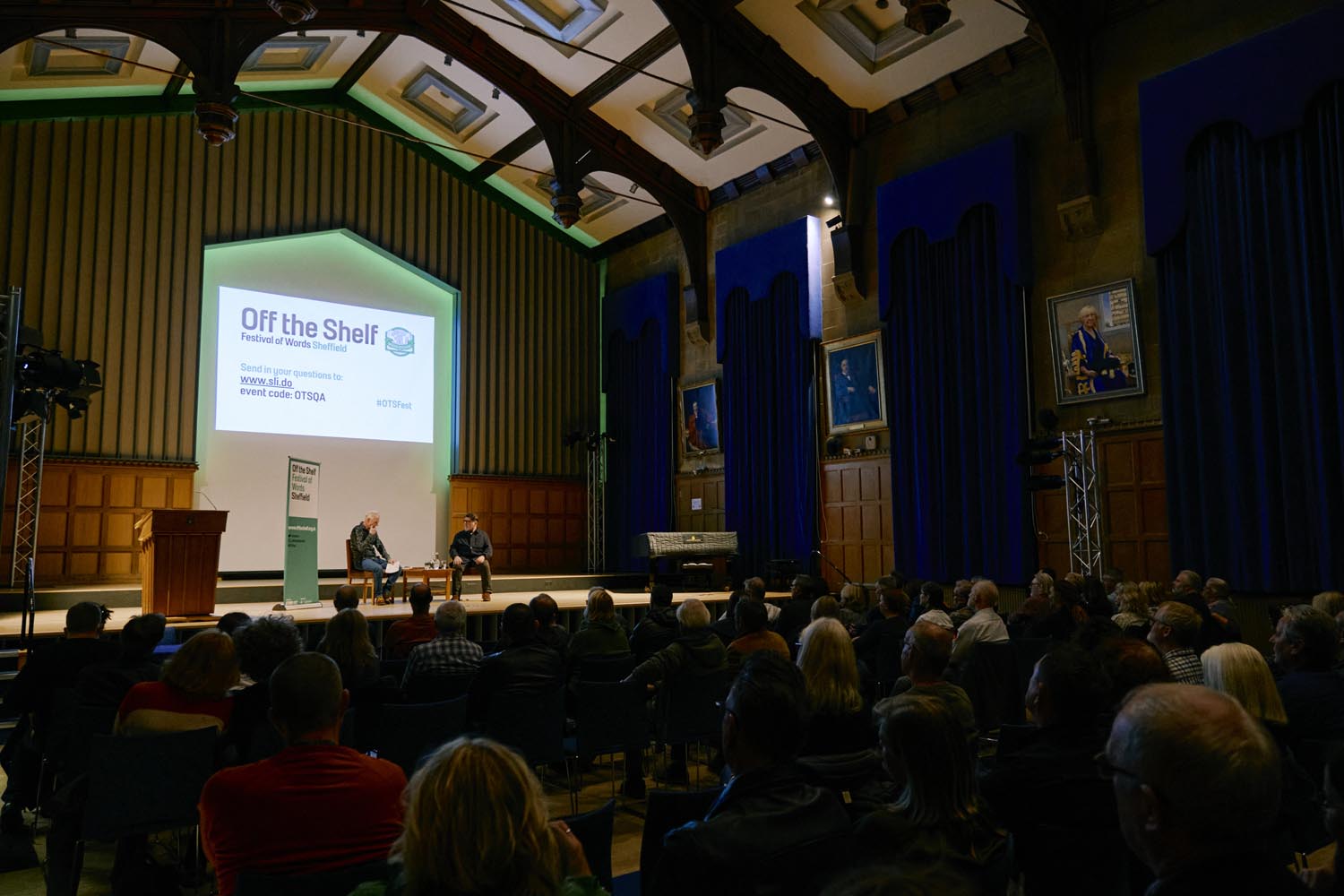 Audience seated in a large hall with high ceilings and blue curtains, watching two speakers on stage during an ‘Off the Shelf’ literary event. A large screen behind the speakers displays event details.