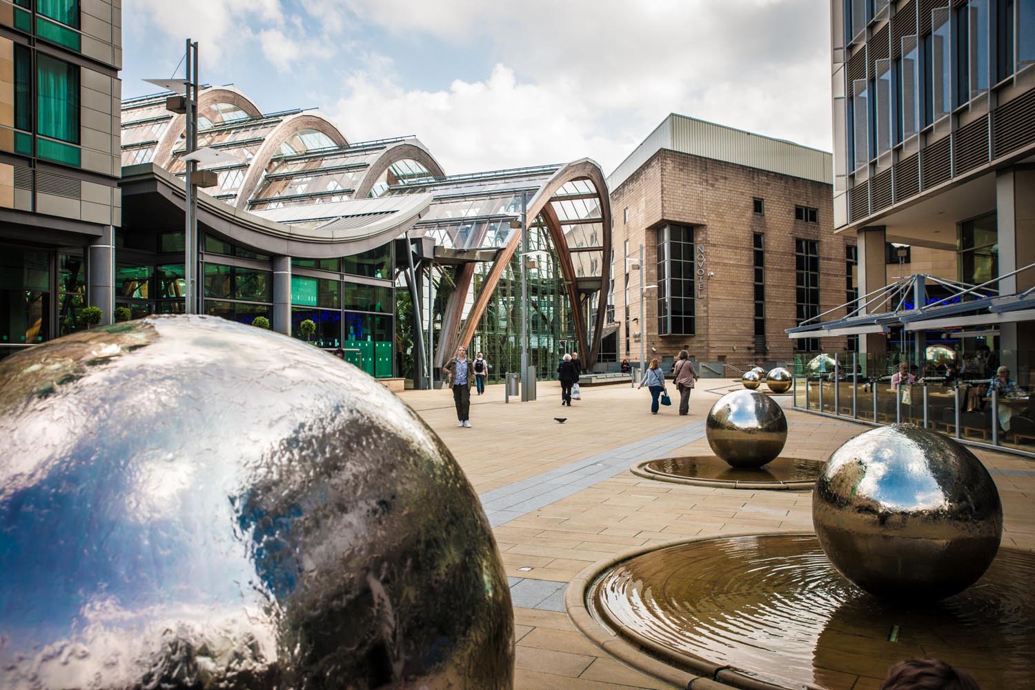 A view across Millennium Square in Sheffield city centre. There are several water features in the shape of large stainless steel balls with water cascading down them. In the background you can see one end of the Sheffield Winter Garden.