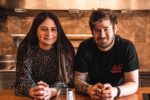Two people sitting at a wooden counter in a modern kitchen setting. One person is wearing a patterned long-sleeve top, and the other is wearing a black T-shirt with a red logo and has visible tattoos on the forearm. Stainless steel surfaces and kitchen equipment are visible in the background.