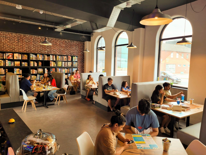 Inside The Dice Box Sheffield, there are tables and booths full of people playing games and drink and eating. The back wall is covered in shelving that is full of boxed games.