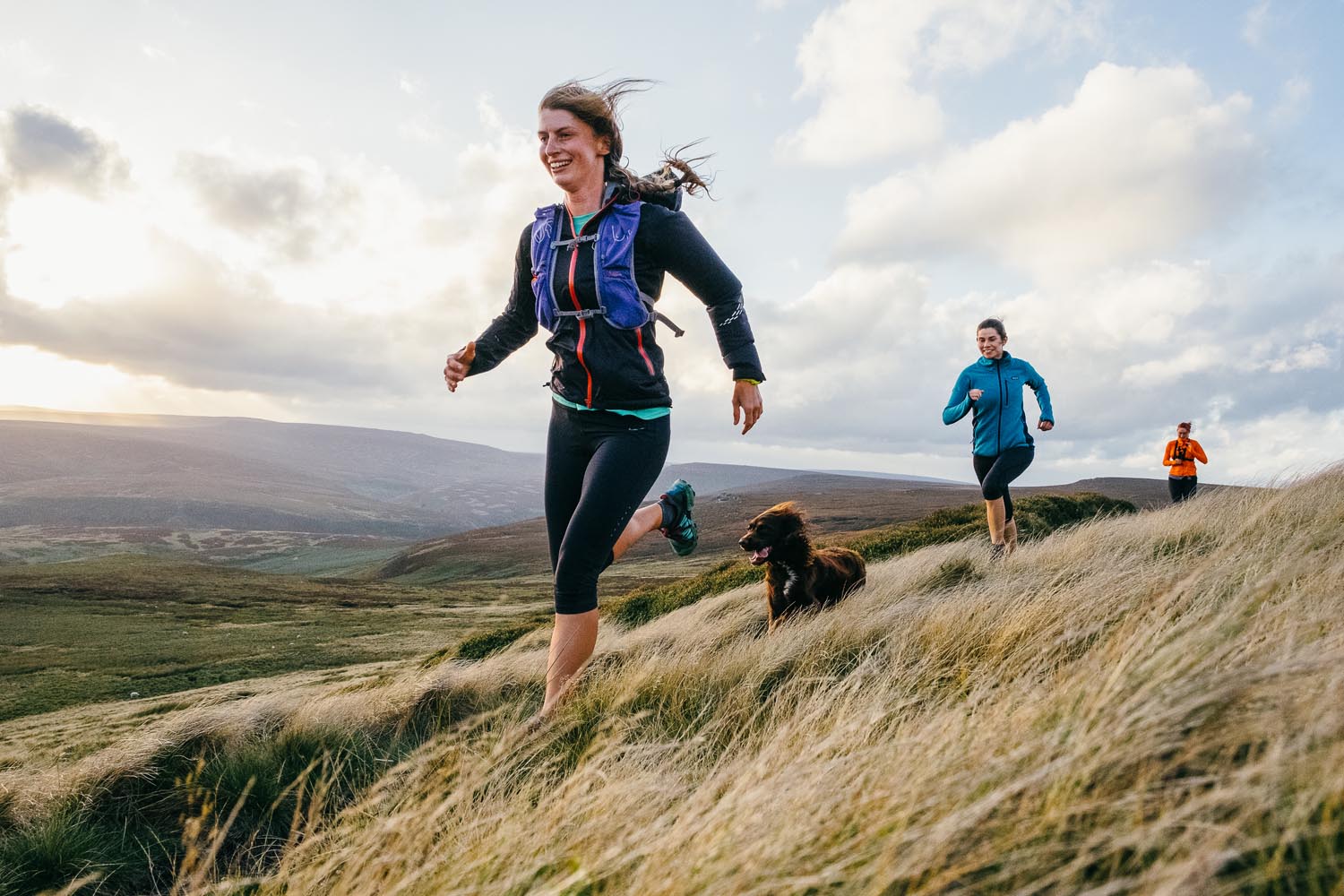 Three people are running in the countryside with their dog.