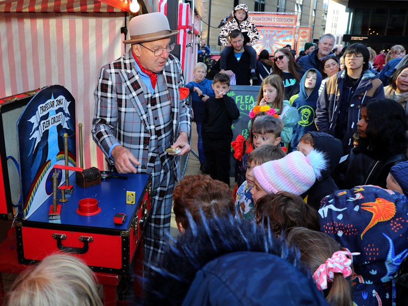 A flea circus themed magician dressed in pinstripe suit and tombola hat entertains a group of children with magic tricks