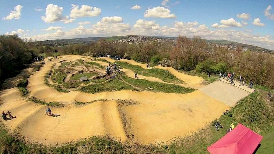 erial view of a dirt bike pump track with winding paths and raised berms surrounded by trees and greenery. Several cyclists are riding on the track, and a red canopy tent is visible in the bottom right corner. The background shows a hilly landscape under a blue sky with scattered clouds.