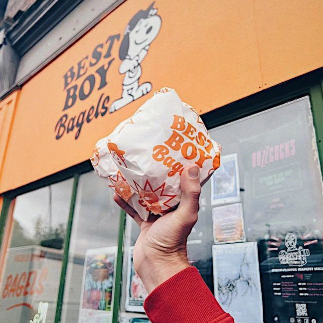 A hand holds a wrapped bagel from Best Boy Bagels, whose sign can be seen in the background.