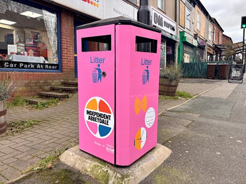 Bright pink litter bin on a pavement with text reading ‘Independent Abbeydale’ inside a circular logo with colorful segments. The bin also features small graphic icons and the word ‘Litter’ near the openings. Shops and a street are visible in the background.