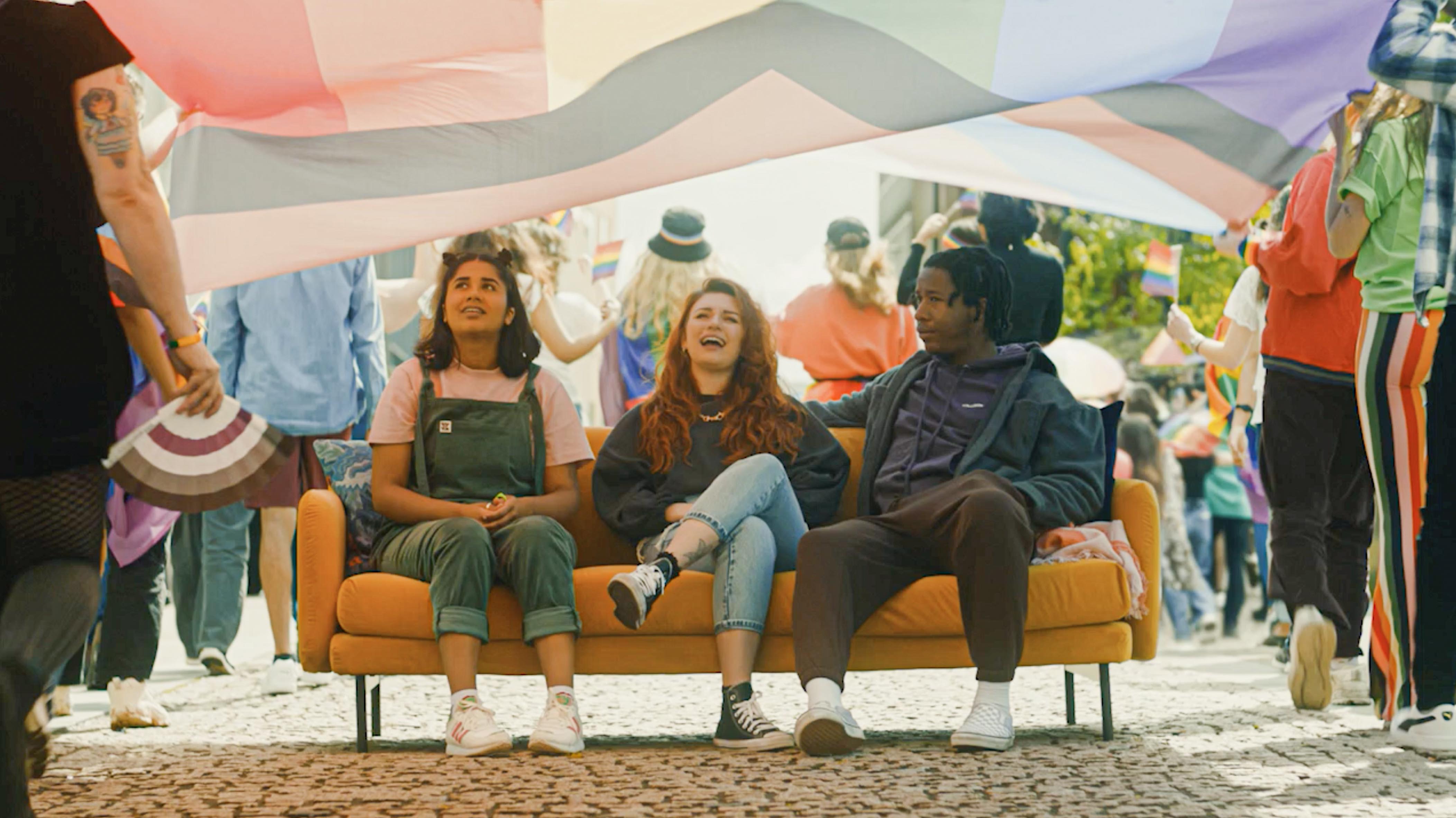 Three people sitting on an orange couch placed outdoors during a parade, with a large rainbow-colored flag held above them. The background shows a crowd of people walking and holding smaller pride flags, creating a festive and colourful atmosphere on a cobblestone street.
