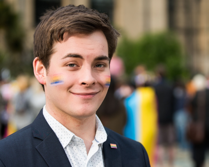 A person wearing a dark blazer over a white patterned shirt is standing outdoors in front of a blurred crowd and historic building. A small rectangular pin with horizontal stripes is attached to the blazer lapel. The background includes greenery and people, some holding colorful flags.