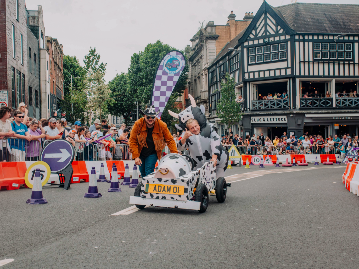 A soapbox cart designed to look like a cow, complete with black-and-white spots and a cow head on the front, is being pushed by a person wearing an orange jacket. Another participant in a cow costume sits inside the cart, raising one arm. The cart has a yellow plate with the text “ADAM 01.” The race takes place on a city street lined with safety barriers, purple-and-white cones, and directional signs, with a large crowd of spectators behind metal railings. 