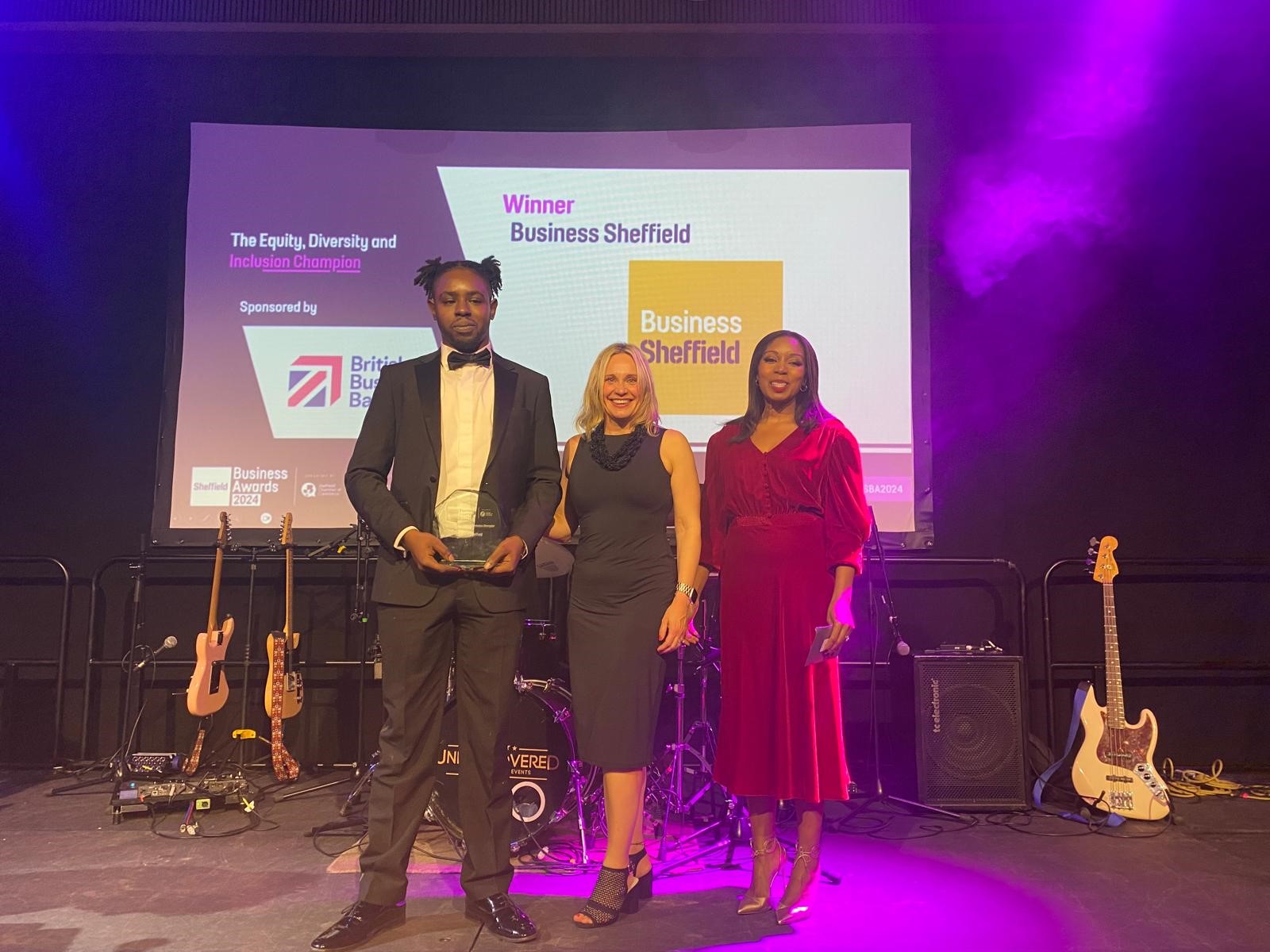 A group of people dressed in formal attire posing together at an indoor event with blue and purple stage lighting. Behind them is a stage with musical instruments and a large screen displaying “Sheffield Business Awards 2023” along with sponsor logos. One person in the center is holding an award, and another on the right is holding a framed certificate. The setting suggests a celebratory awards ceremony.