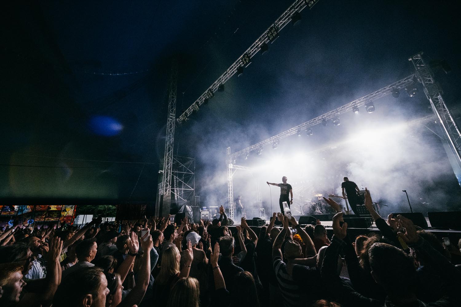 A lively concert scene with a large crowd raising hands toward a brightly lit stage. Two performers stand under spotlights amid smoke effects, with tall metal rigging and speakers framing the stage. The dark canopy above contrasts with the intense white stage lights, creating a dramatic atmosphere.