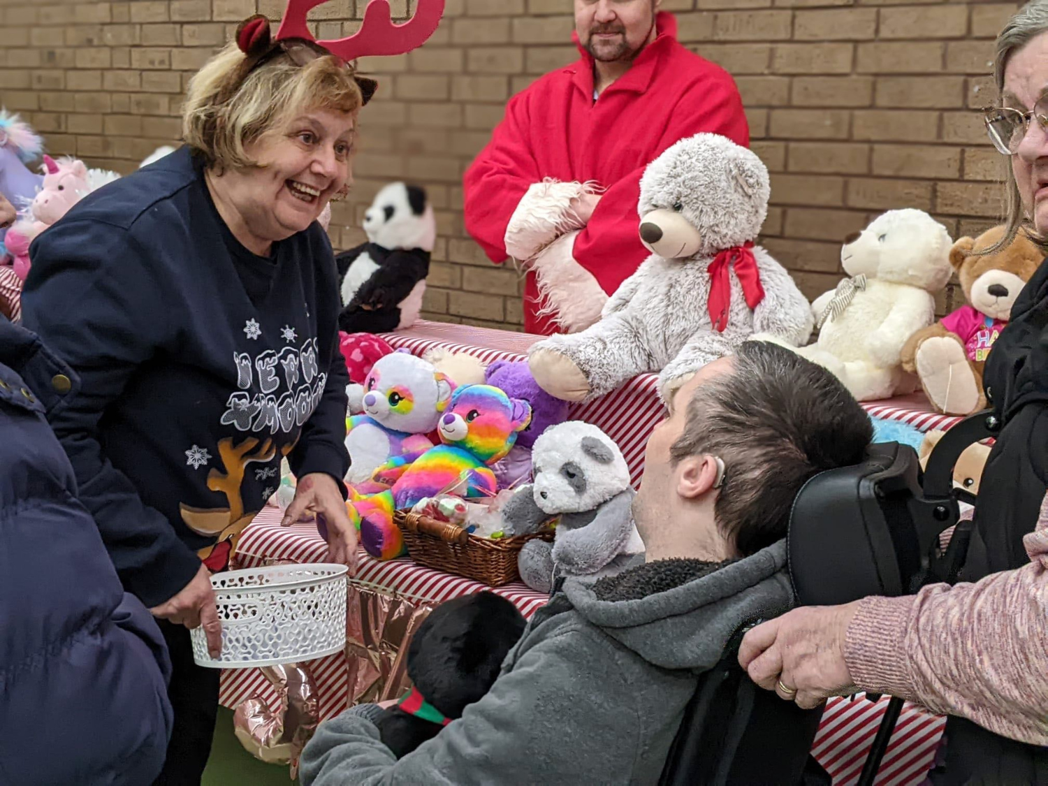 Indoor Christmas market stall with a table covered in red-and-white striped cloth displaying plush toys, including teddy bears, pandas, and colorful rainbow animals. A person in festive antlers holds a white basket while interacting with another person in a wheelchair. Additional people stand nearby, and the background features a brick wall.