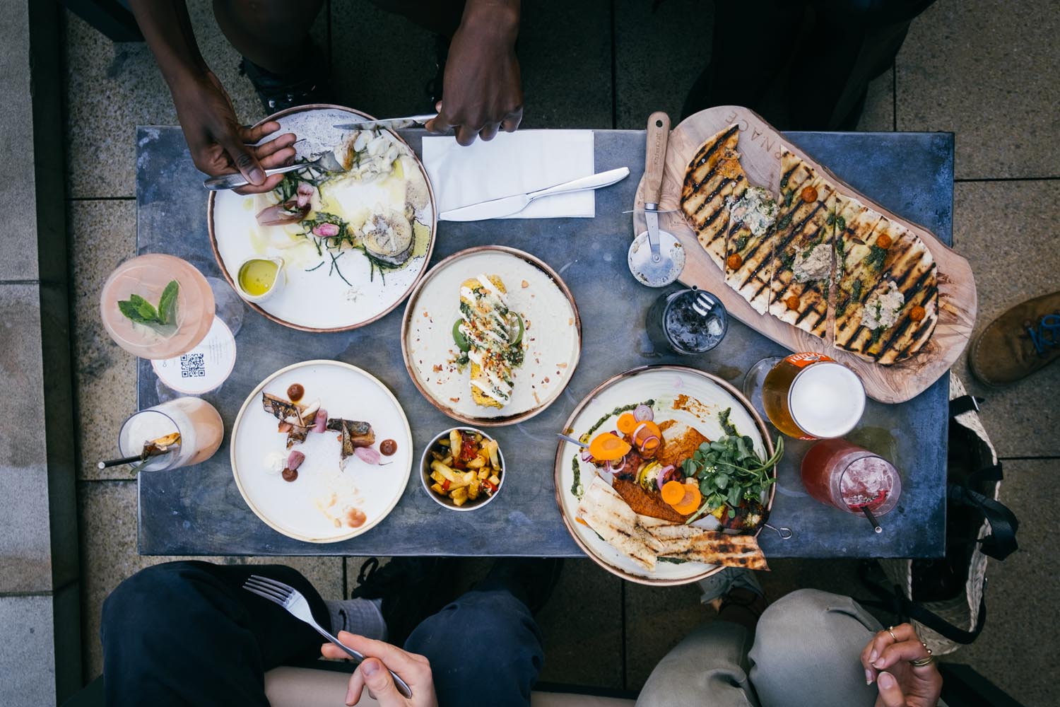 An overhead shot of a table in a restaurant filled with plates of food and drinks. There are people, mostly out of shot, enjoying the meal.