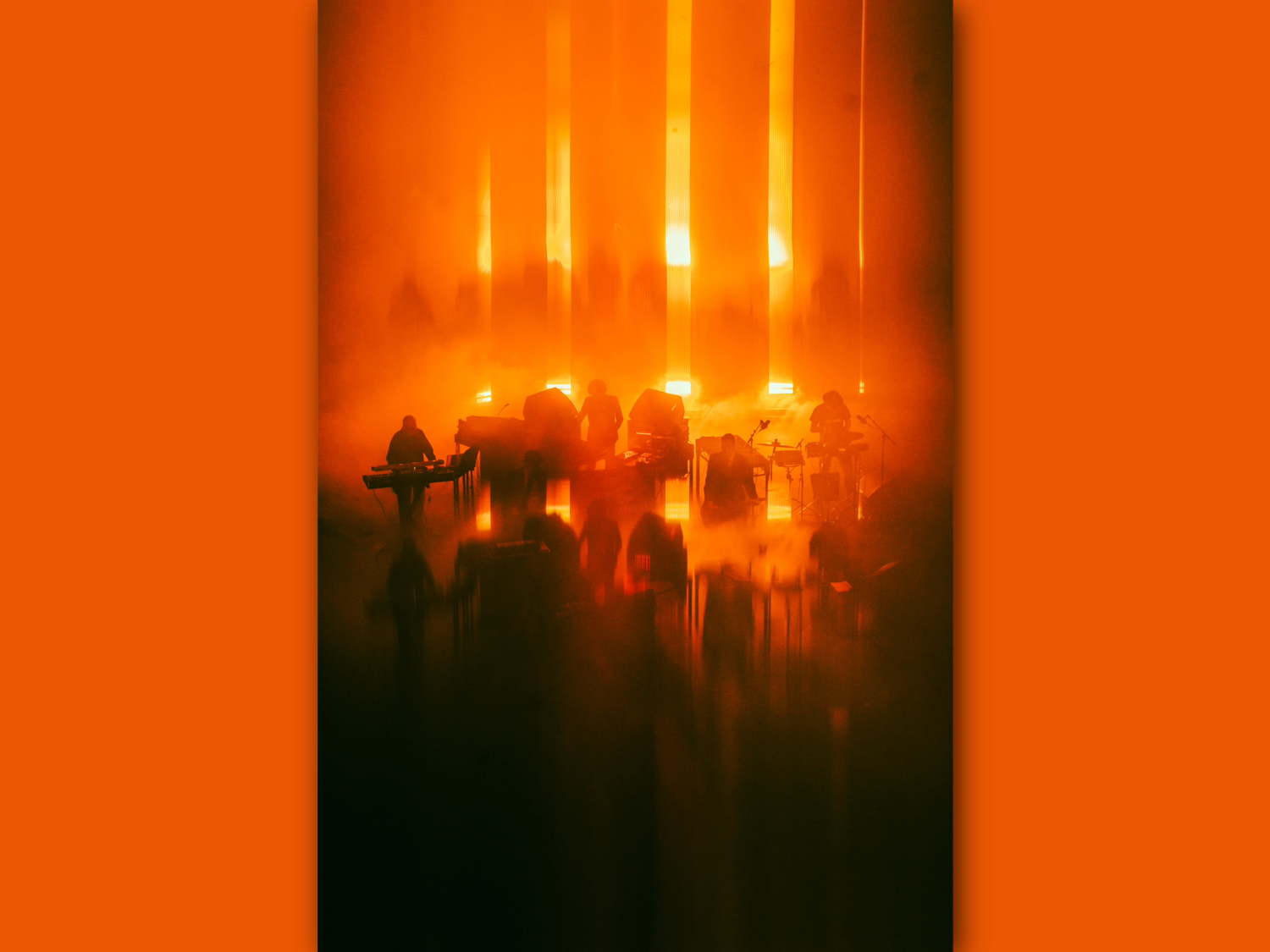 Silhouetted musicians perform on stage surrounded by dense orange fog and dramatic vertical light beams. The scene is bathed in warm, glowing tones, creating an abstract, atmospheric effect with blurred reflections on the floor.