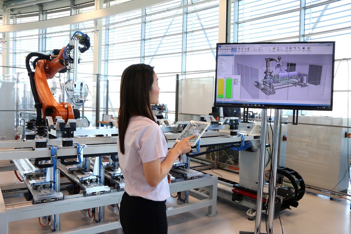 A woman is standing in front of a monitor and holding a tablet. In the background is an industrial robot arm.